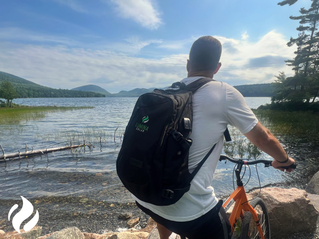 Man with backpack standing by a bike, looking out over a lake surrounded by mountains.